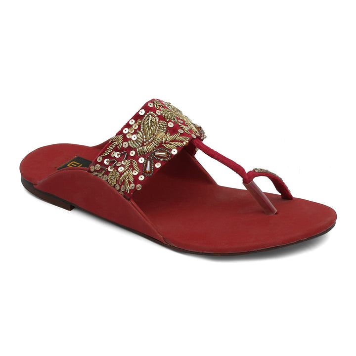 A red flat sandal with traditional Kolhapuri embellishments, featuring a toe loop and a decorative strap with metal and bead details.