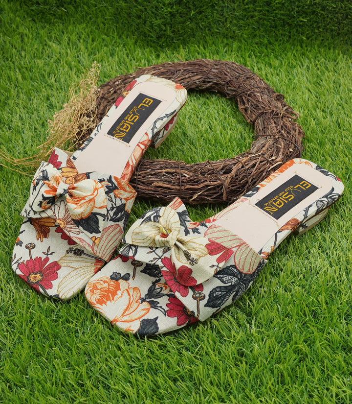 A pair of multicolored floral slipper socks with a bow on the top, displayed on a grassy surface next to a decorative woven ring.