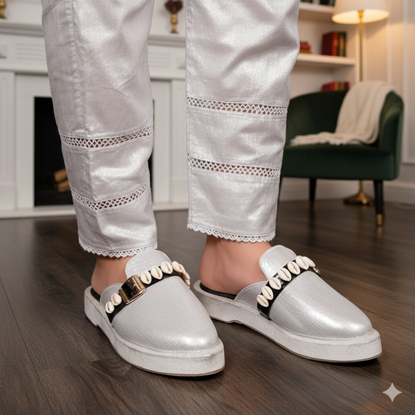 Person wearing silver slip-on shoes with decorative straps in a room setting.