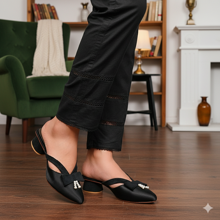 Person wearing black heeled shoes with a bow in a room with a green chair and bookshelf.