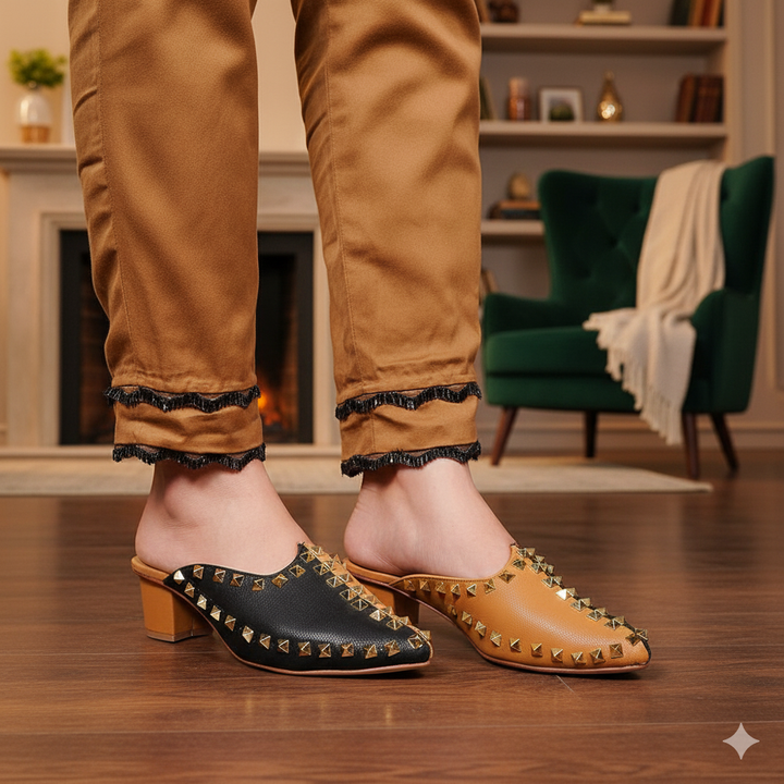 Person wearing tan pants and black and gold studded shoes in a room with green chairs and a fireplace.